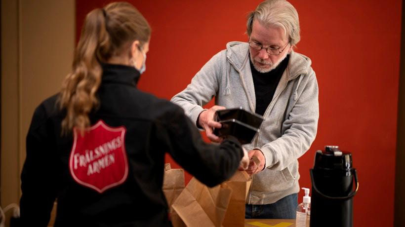 En kvinna från Frälsningsarmén med ryggtavlan mot kameran. Hon delar ut en matkasse med matlådor till en äldre man som står mitt emot henne.