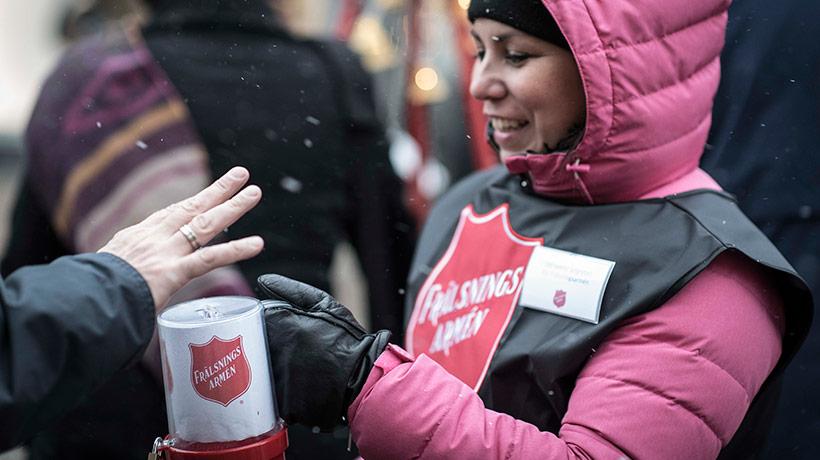 En leende kvinna i vinterkläder håller i en insamlingsbössa - en hand syns som stoppar i pengar i bössan.