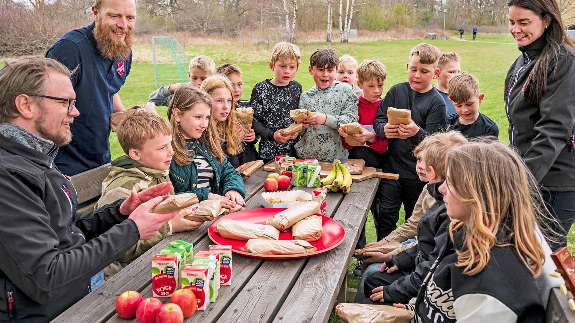 Ett gäng barn runt ett picknickbord en sommardag tillsammans med två tre vuxna personer som har Frälsningsarméns sköld på tröjan.