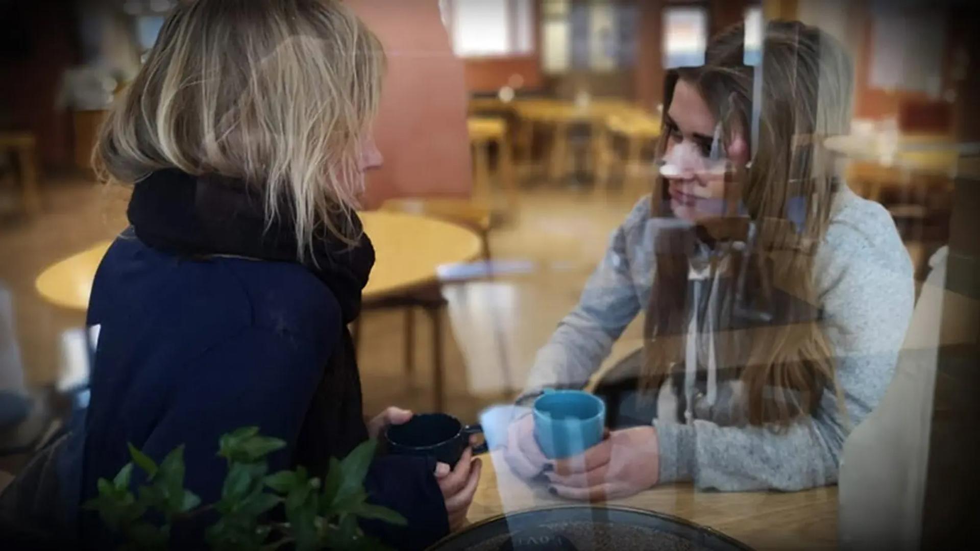 Två kvinnor sitter innanför ett fönster vid ett bord med varsin mugg i händerna.