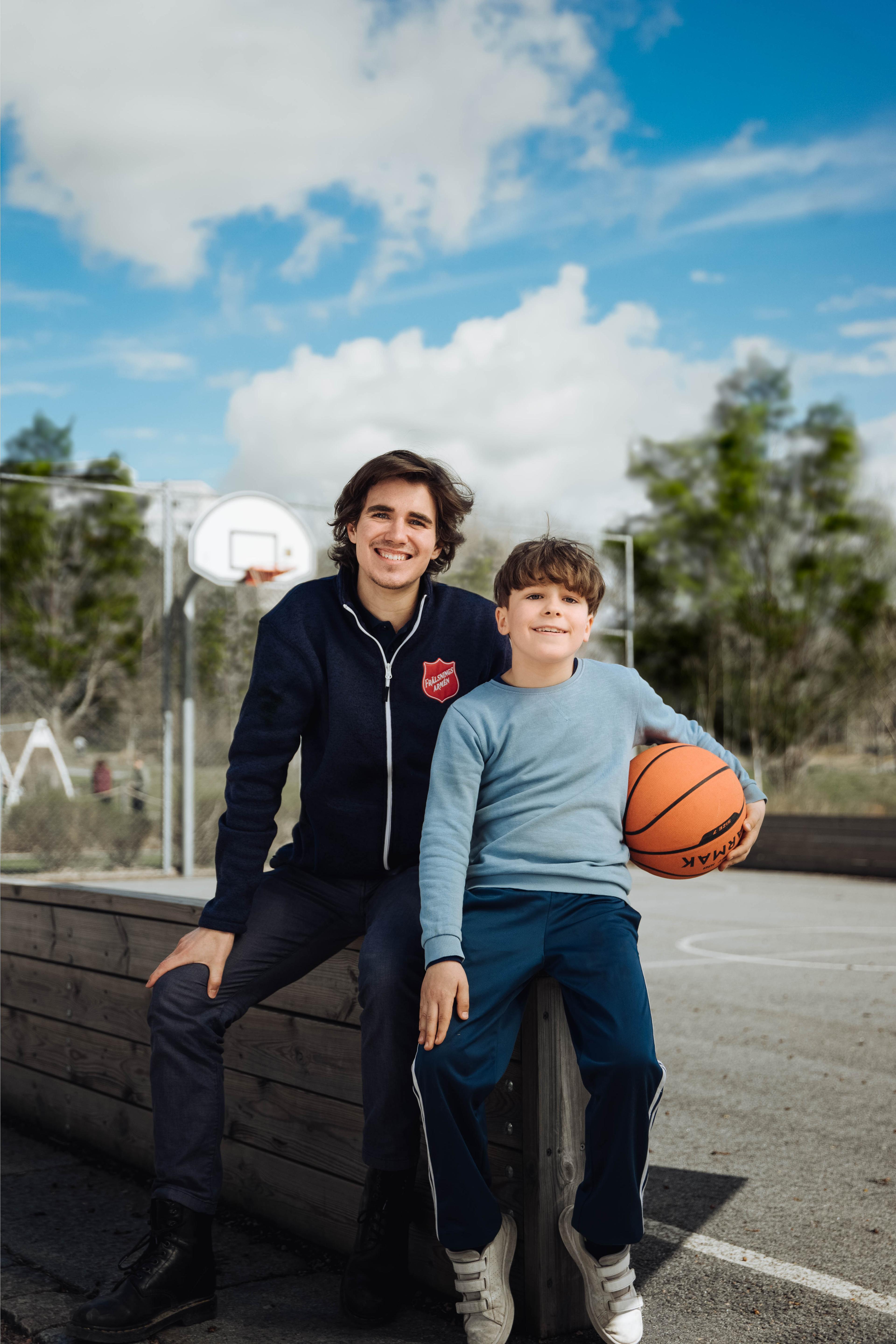 En yngre man med Frälsningsarméns sköld på jackan sitter på en bänk vid en basketplan med en liten pojke som håller i en basketboll. Båda ler in i kameran.