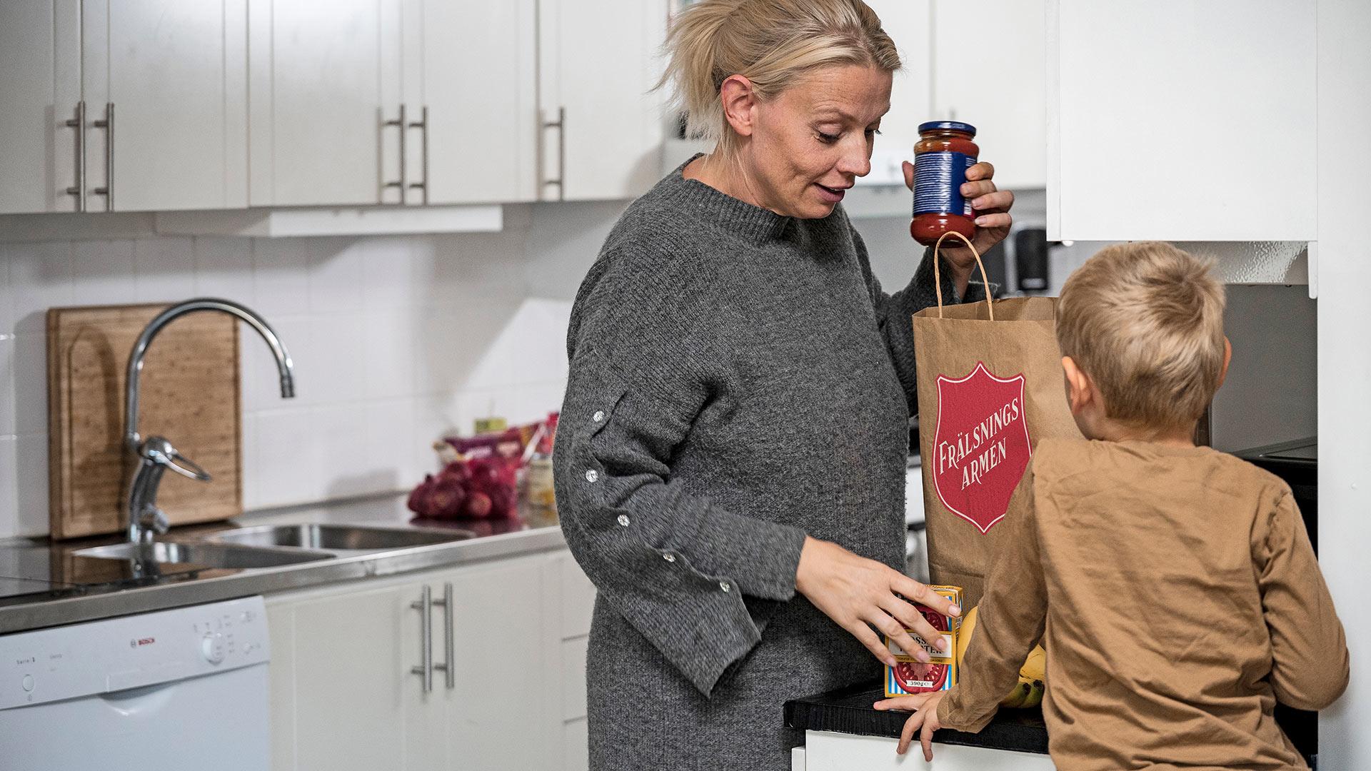 En mamma står i ett kök och plockar upp matvaror från en matkasse med Frälsningsarméns sköld på. En liten pojke syns i förgrunden.