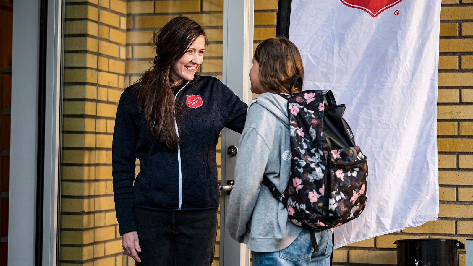 En kvinna med Frälsningsarméns sköld på jackan står i en dörröppning och tar leende emot en flicka med ryggsäck.