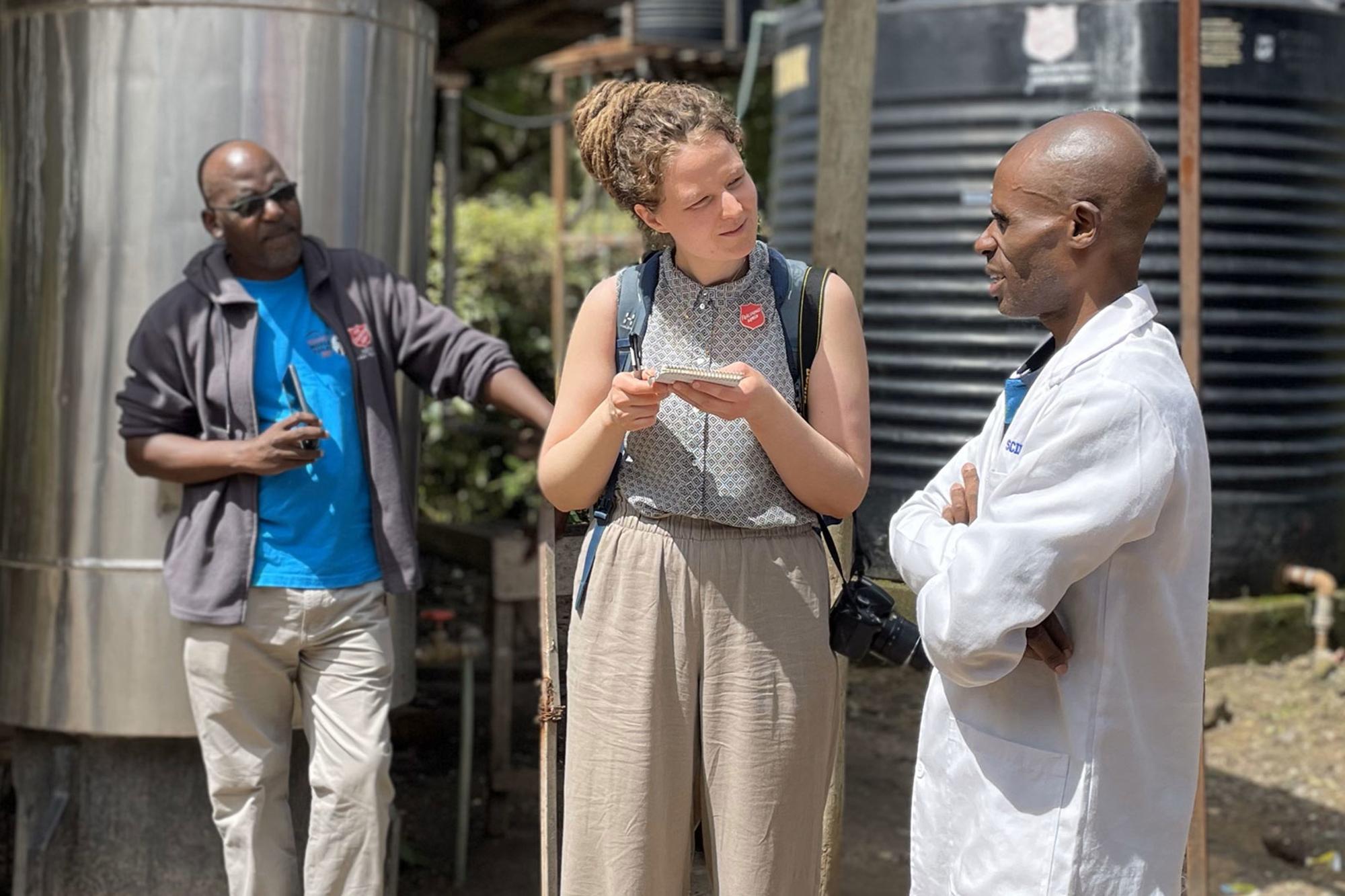 Tre personer med Frälsningsarméns sköld på kläderna står och pratar framför några vattentankar i en by i Kenya.