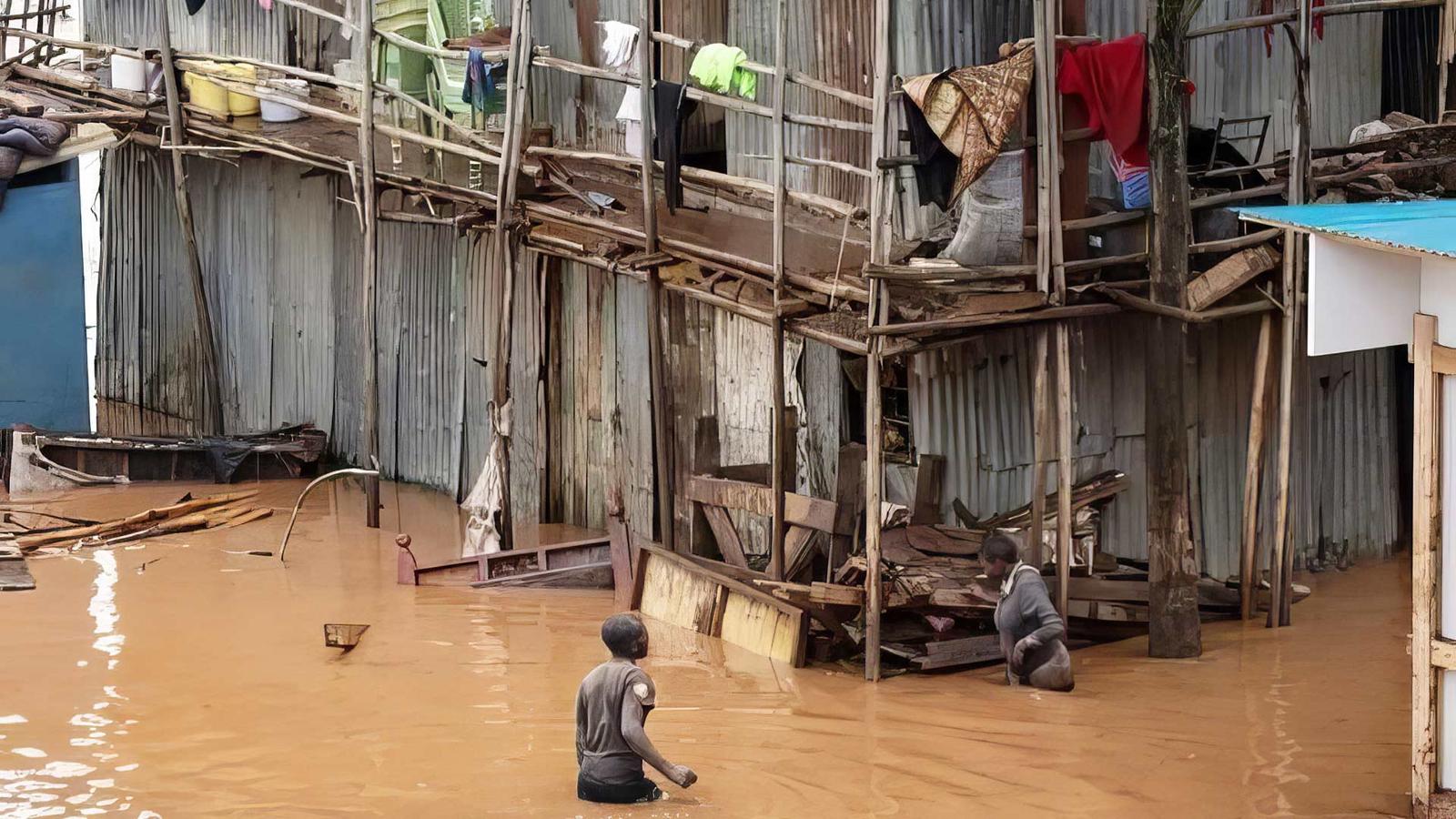 Översvämning i Kenya - två personer vadar i vatten som går upp till midjan, bakom dem syns en stor barack med en massa bråte och två våningar och loftgång. 