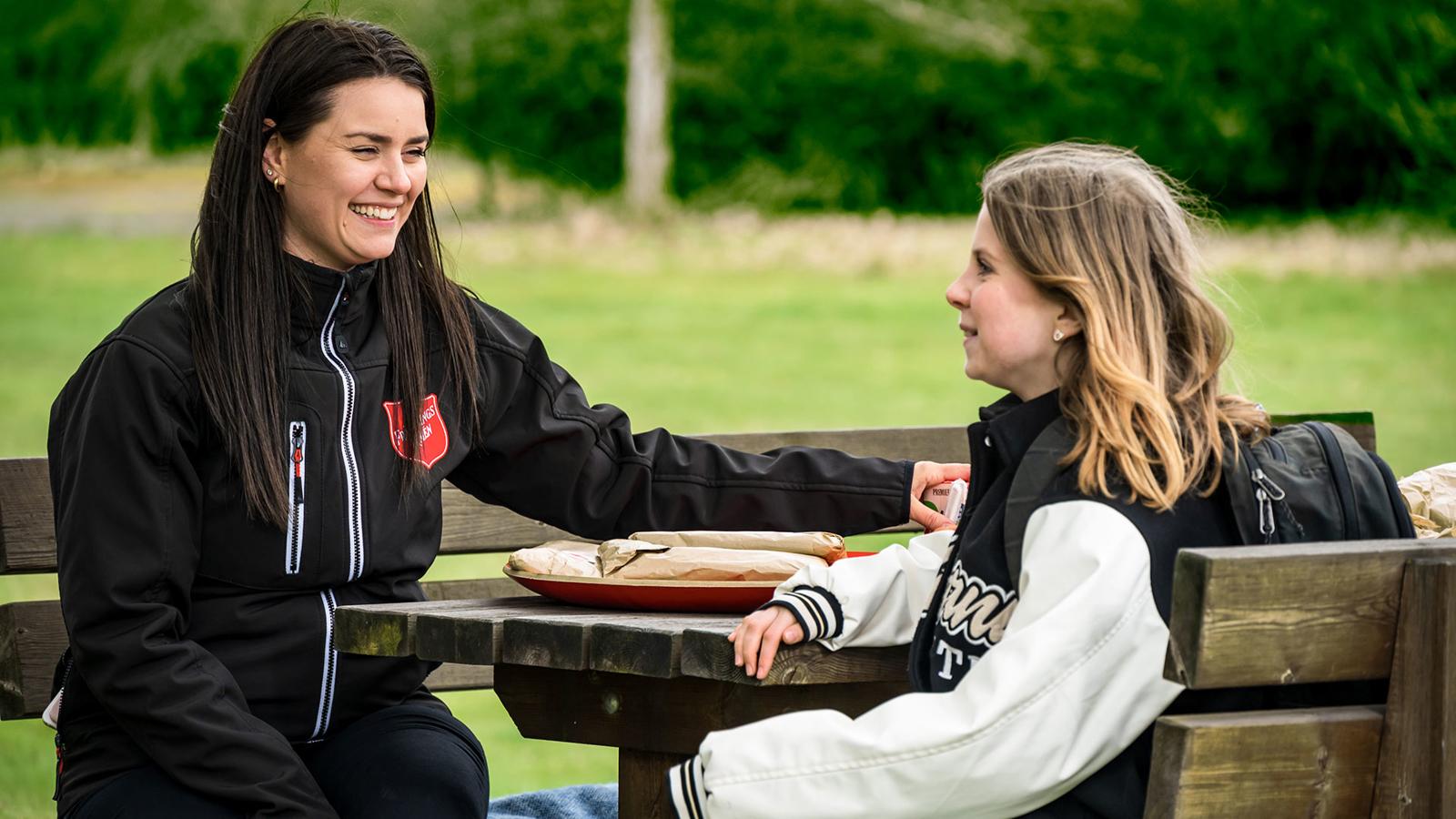 En ung flicka sitter vid ett picknickbord tillsammans med en glad kvinna som har Frälsningsarméns sköld på jackan. I bakgrunden syns ett grönområde.