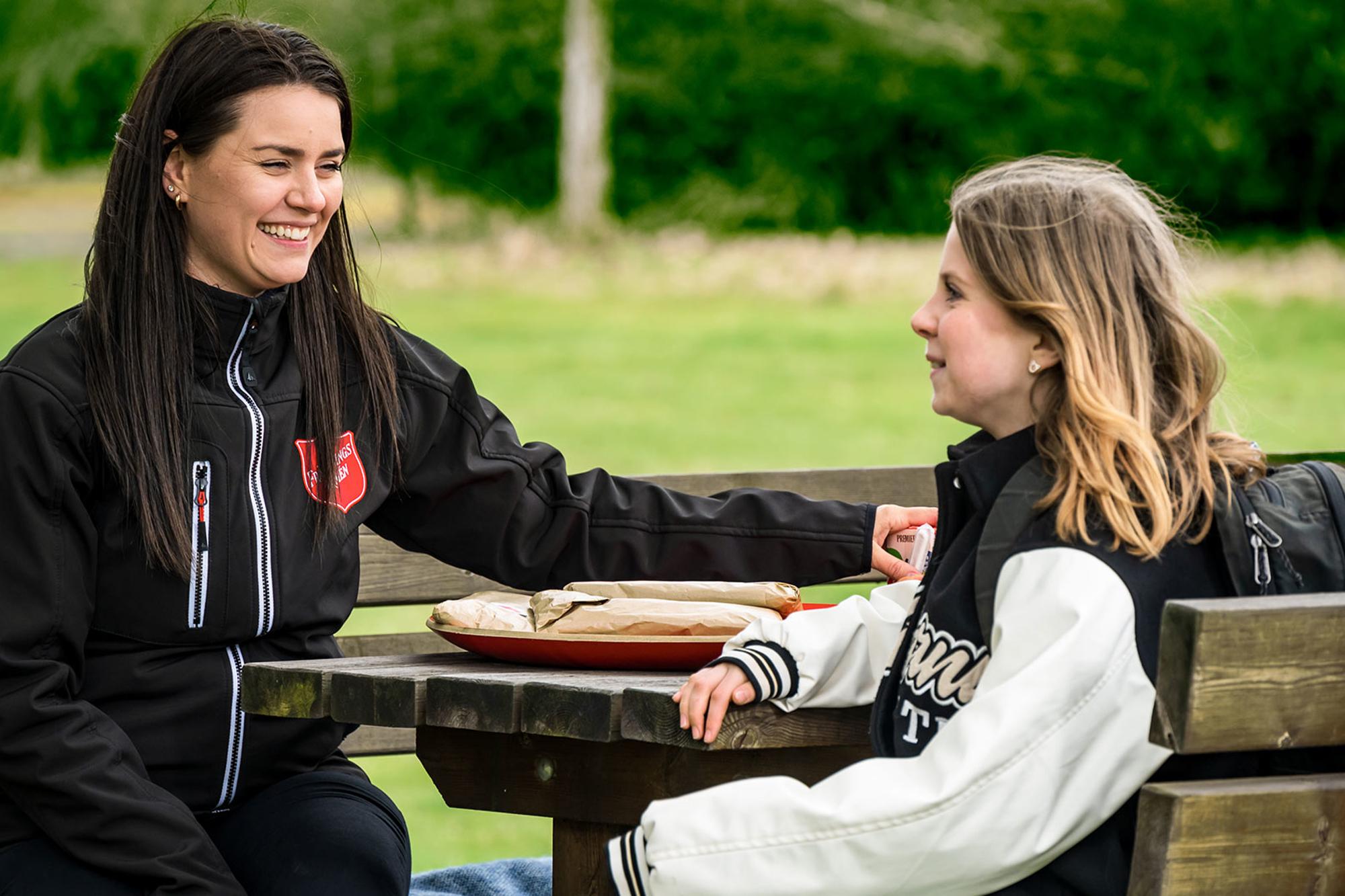 En ung flicka sitter vid ett picknickbord tillsammans med en glad kvinna som har Frälsningsarméns sköld på jackan. I bakgrunden syns ett grönområde.