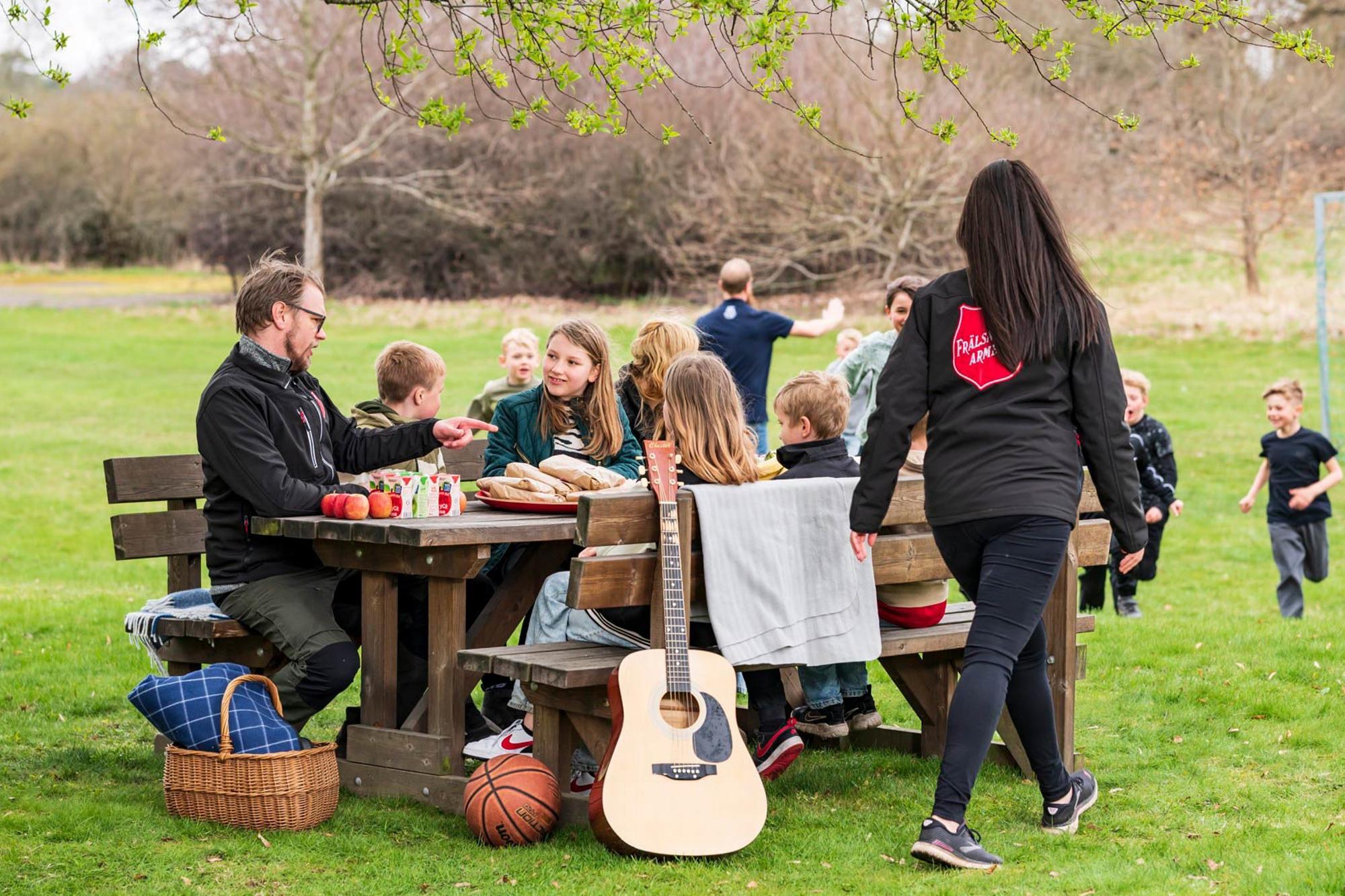 Några barn och vuxna kring ett picknickbord med frukt och juice, på sidan syns en gitarr, en basketboll och en picknickkorg. I bakgrunden syns ett grönområde med barn som spelar fotboll.