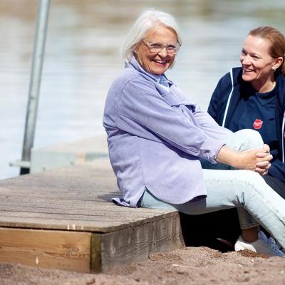 En äldre kvinna sitter leende på en brygga  vid en sjö tillsammans med en yngre kvinna som har Frälsningsarméns sköld på tröjan.