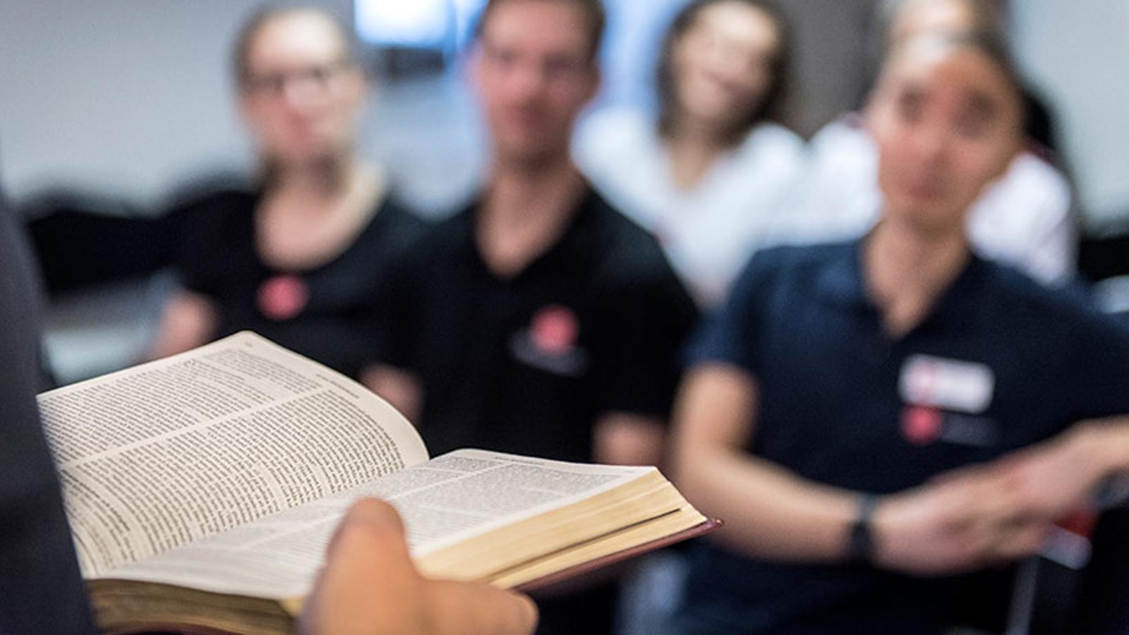 En person som håller i en uppslagen bibel. I bakgrunden står personer från Frälsningsarmén och lyssnar.