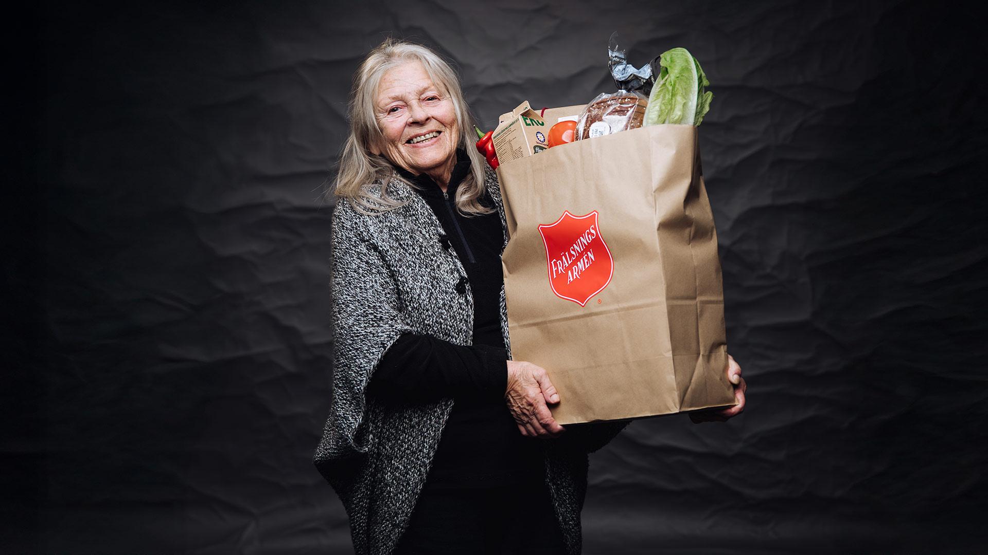 En leende äldre kvinna håller i en full matkasse i famnen.