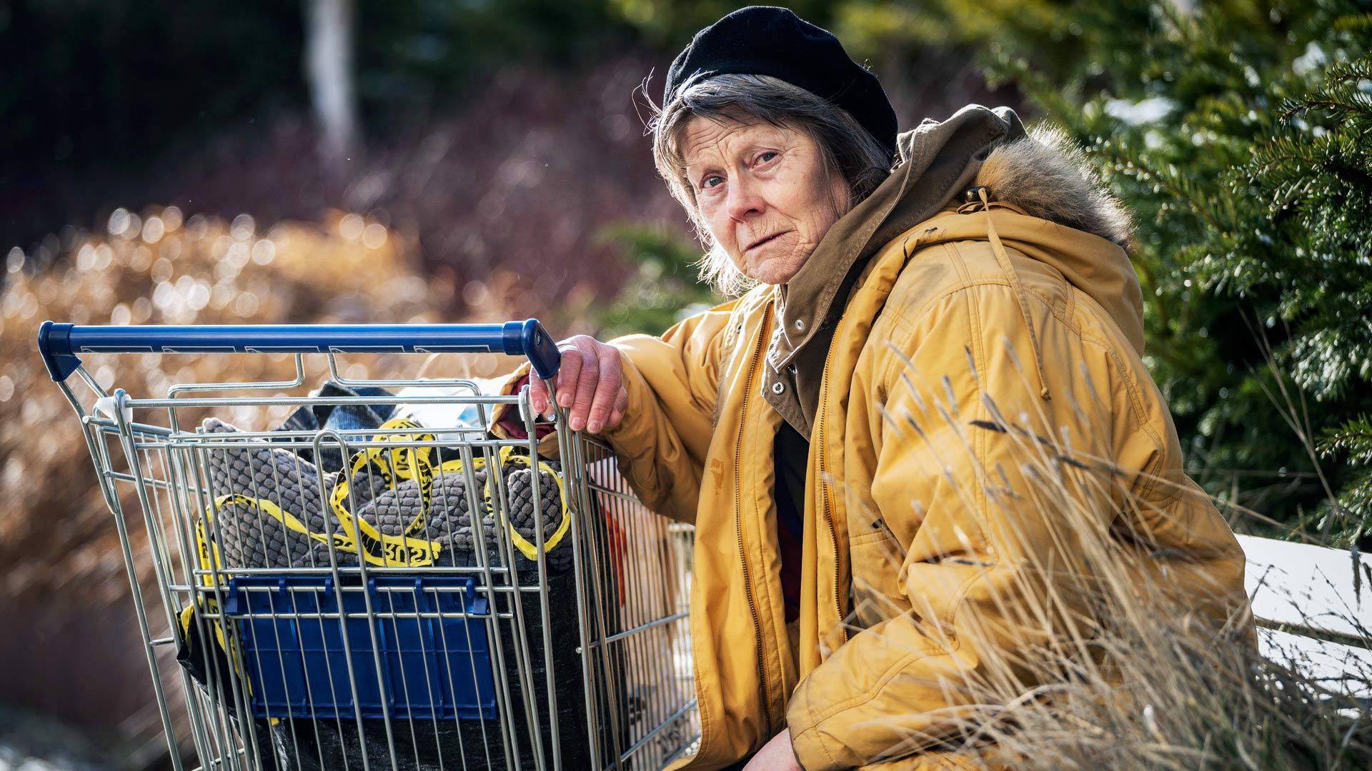 En äldre kvinna med allvarlig blick sitter på en parkbänk med en kundvagn framför sig med påsar och väskor i. I bakgrunden en parkmiljö