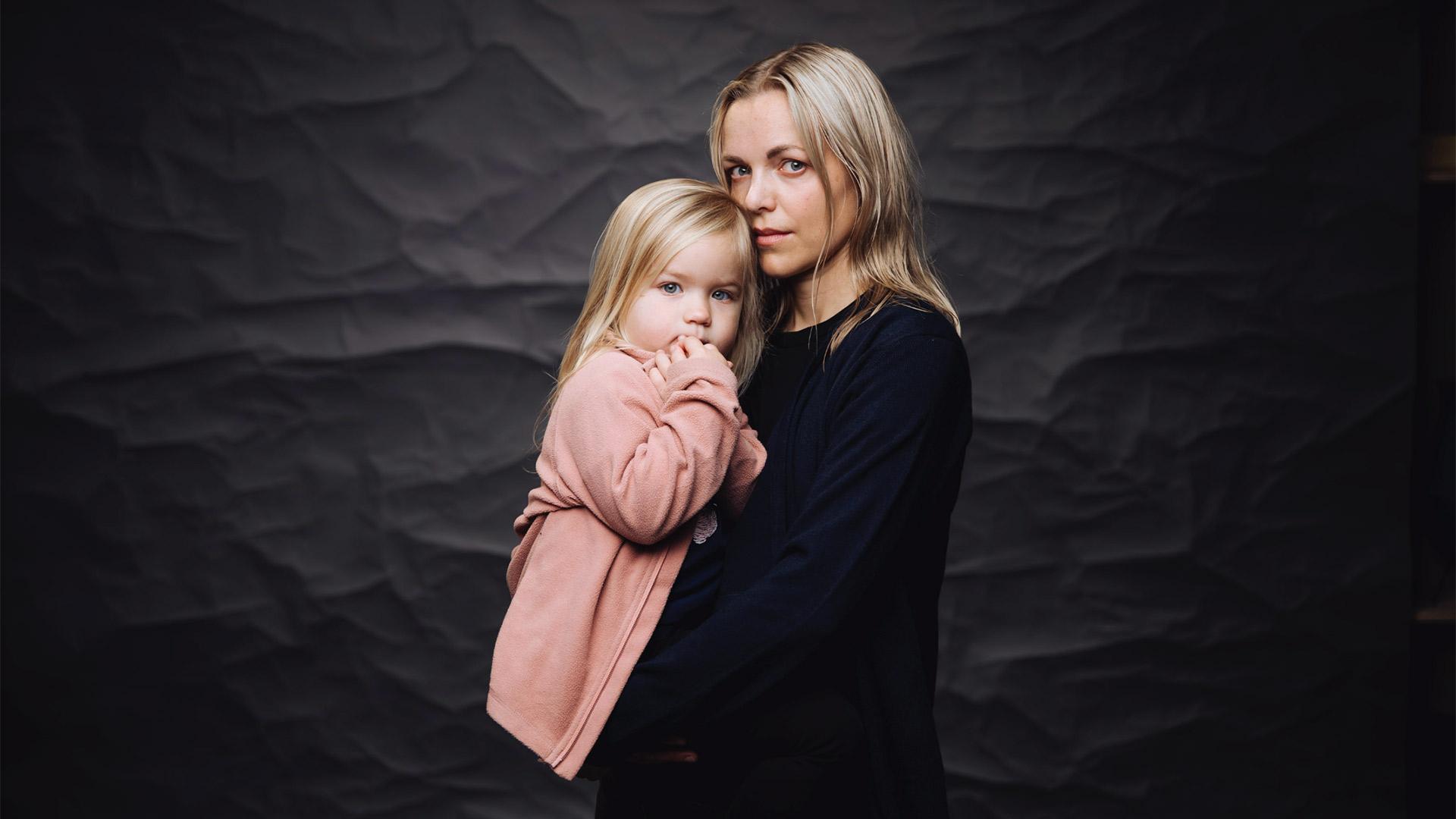 En ung mamma med en allvarsam blick i kameran och en liten dotter i famnen