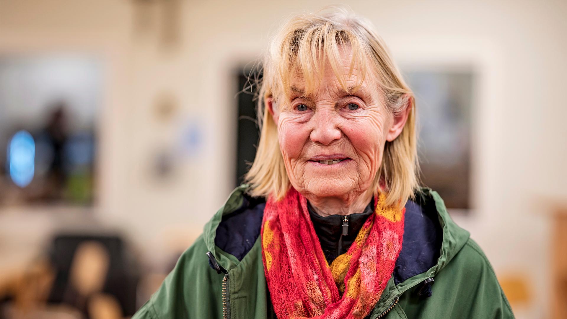 En äldre dam iklädd en grön jacka med en röd scarf runt halsen som tittar in i kameran.