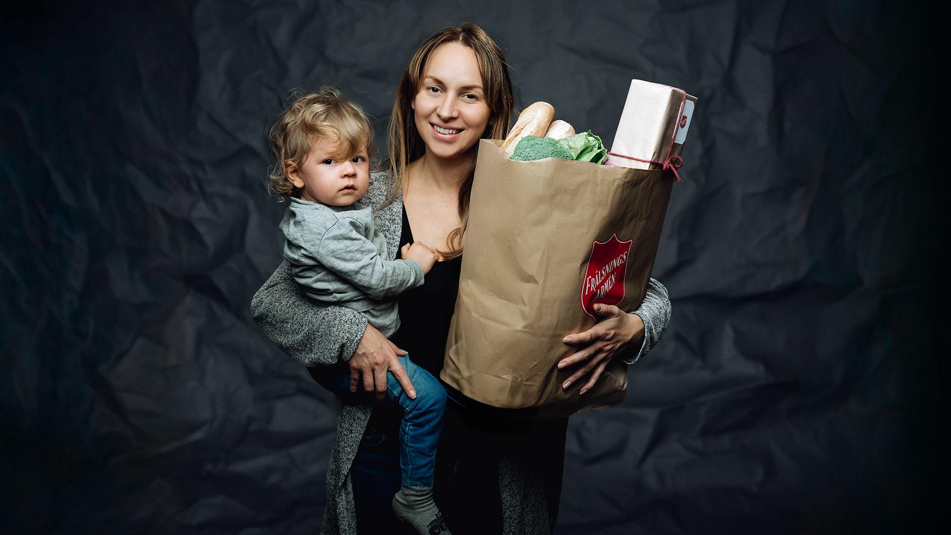 En leende ung kvinna med ett barn på ena armen och en matkasse fylld med mat under andra armen.