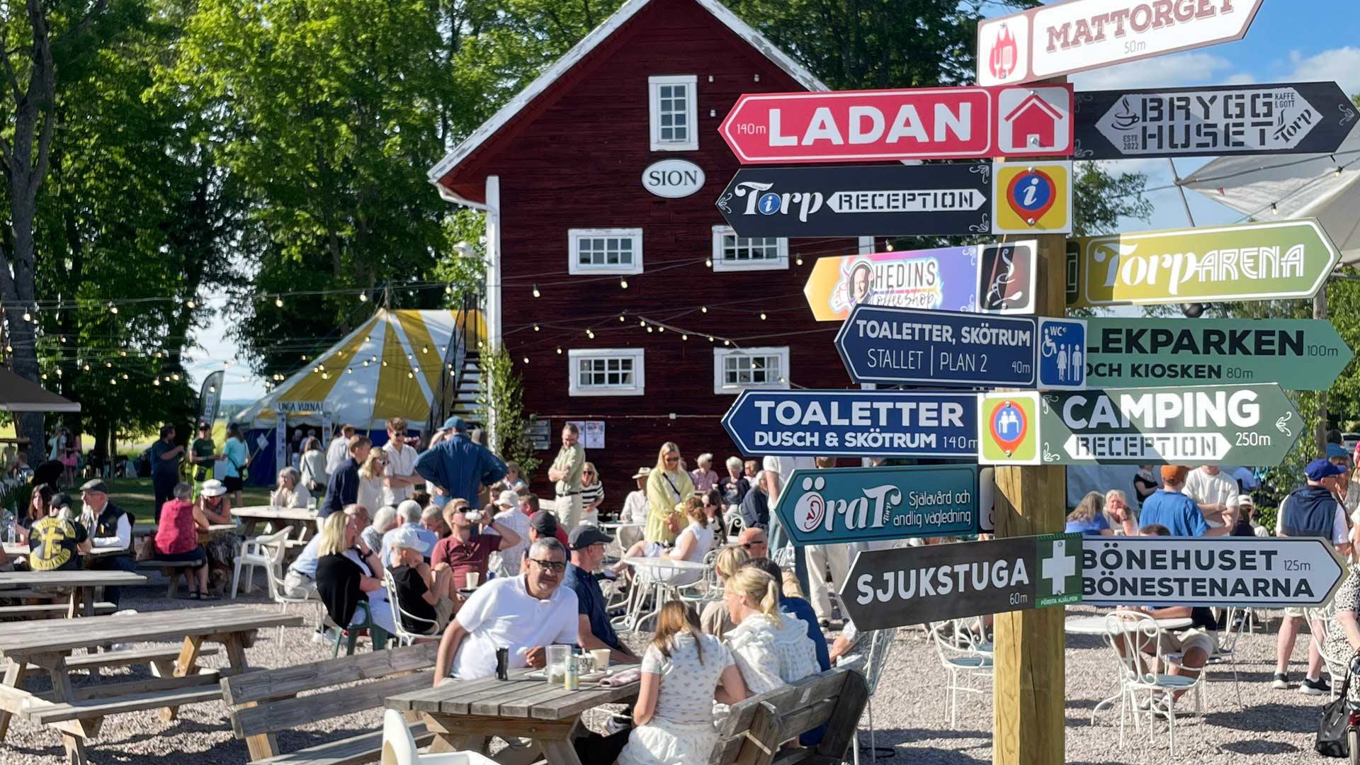 Människor sitter vid picknickbord, solen skiner och i bakgrunden syns träd, ett rött trähus och tält. I förgrunden en stolpe med ett flertal skyltar bland andra Torp Arena.